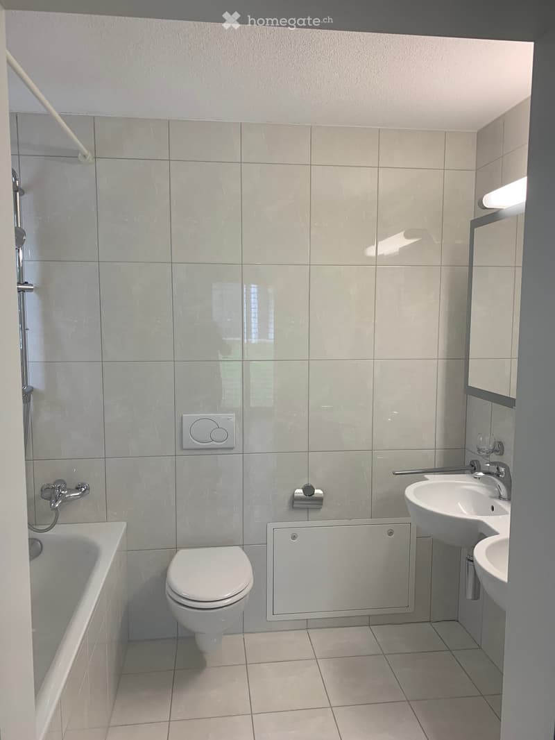 Modern bathroom with white tiles, bathtub, toilet, double sink, and showerhead.