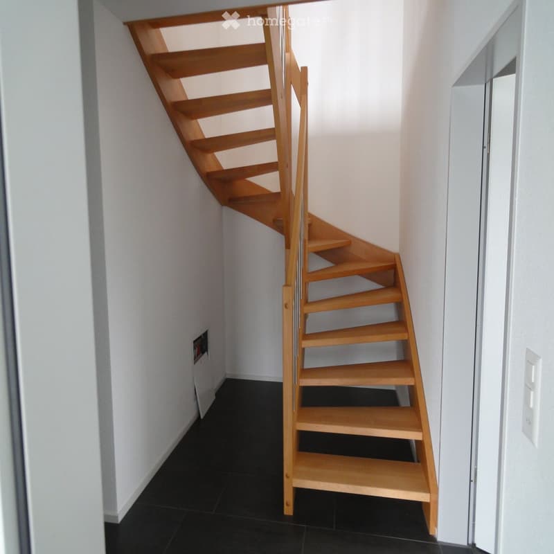 Spiral staircase with wooden steps and handrail, white walls, black tiled floor, and a white door frame on the right side.
