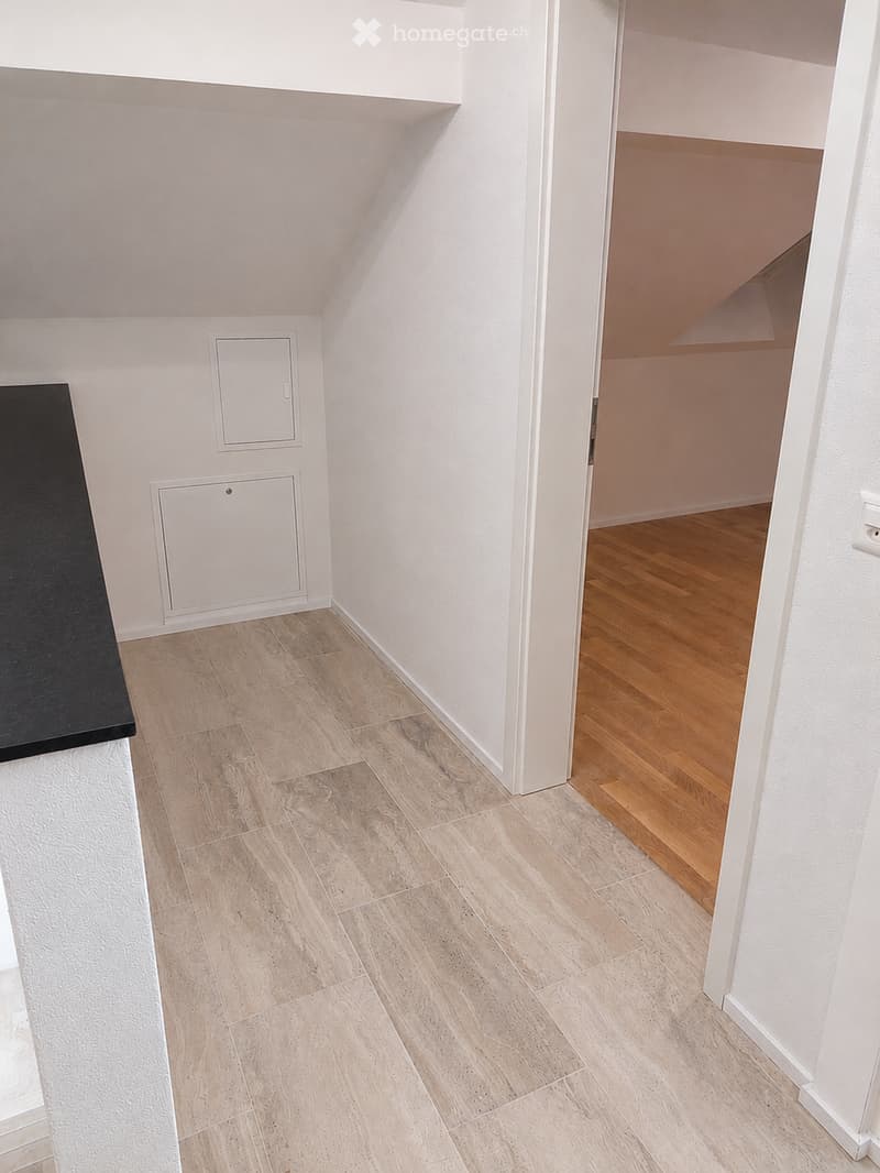 A narrow hallway with light-colored tiled flooring and white walls, leading to a wooden-floored room.