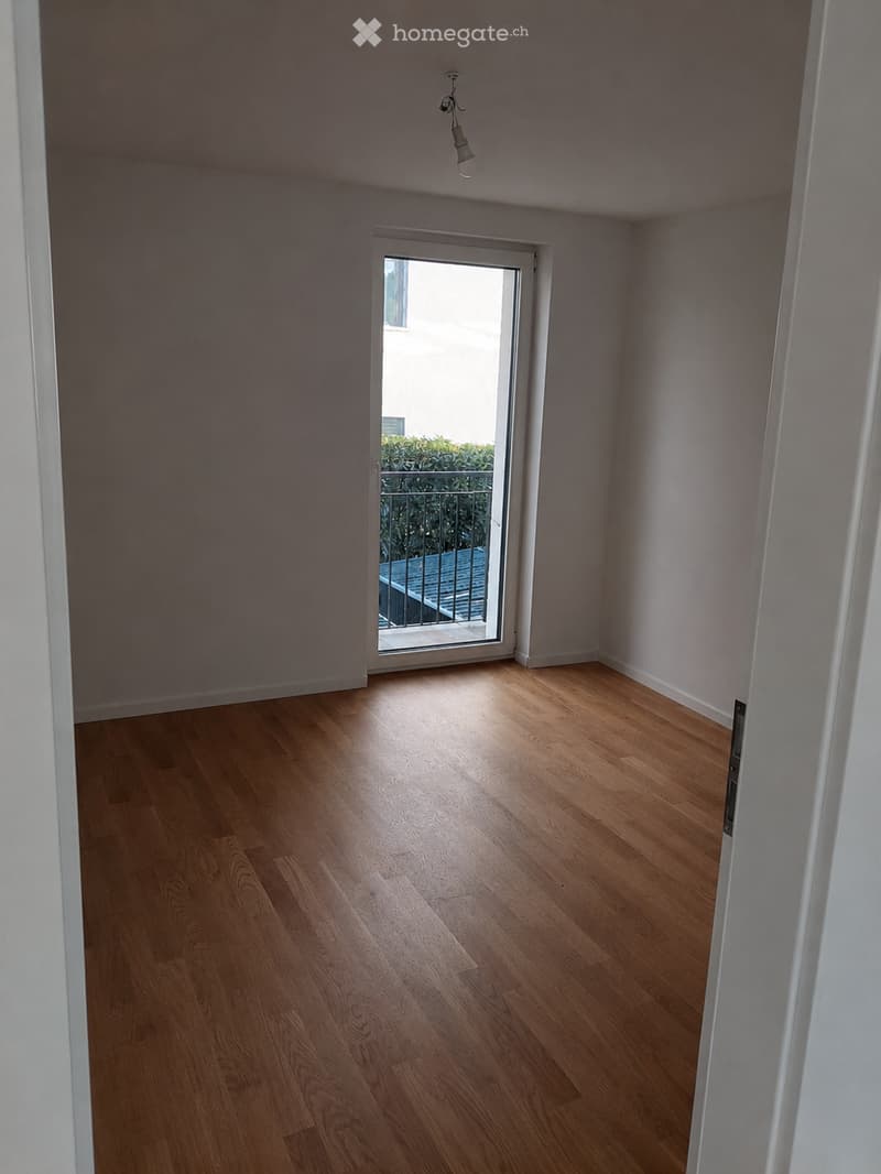 Empty room with light wooden flooring, white walls, and a sliding glass door leading to a balcony with a metal railing and greenery visible outside.