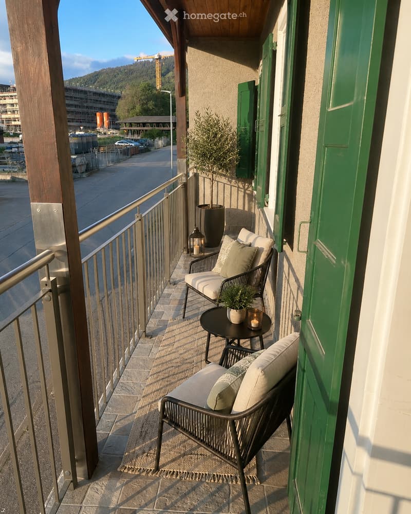 Balcony with two wicker chairs, a small round table, a potted plant, and a candle, overlooking a street with a view of a construction site and mountains.