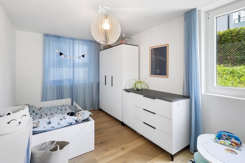 A modern, well-lit bedroom with a white bed, a wardrobe, a nightstand, a small table, and a chair. The room features light blue curtains, a large window with a view of greenery, and decorative items including a framed picture and a potted plant.