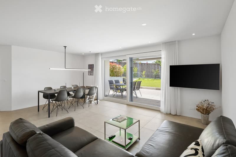 Modern living and dining area with a black leather sofa, glass coffee table, dining table with chairs, and a large TV mounted on the wall. Large sliding glass doors open to a patio with outdoor furniture.