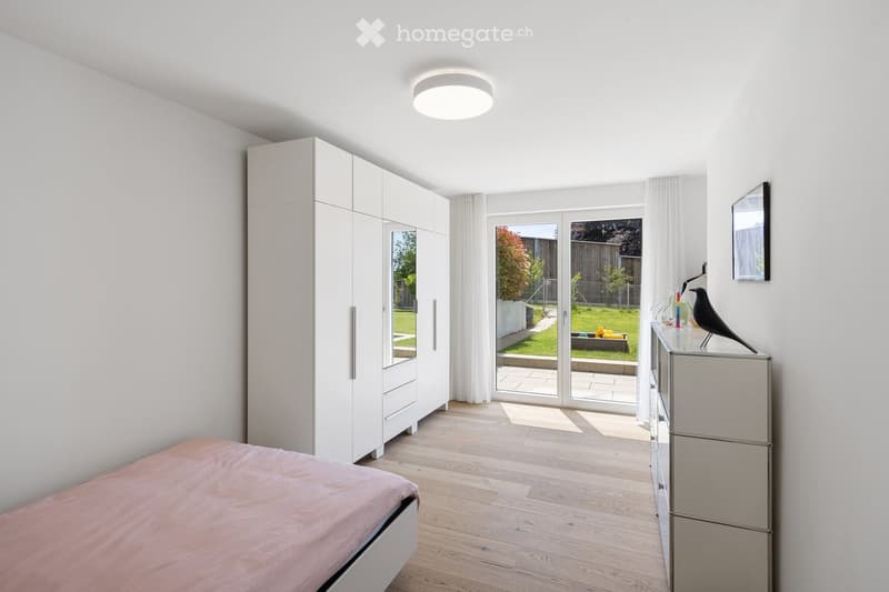 Modern bedroom with light wooden flooring, white walls, a large wardrobe with mirrored doors, a bed with pink bedding, a nightstand with a decorative bird figurine, and large glass doors leading to a garden.