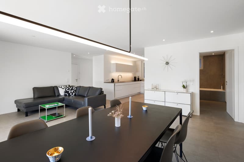 Modern open-plan living and dining area with a black leather sofa, coffee table, dining table with chairs, and a small kitchen area with cabinets and a sink.