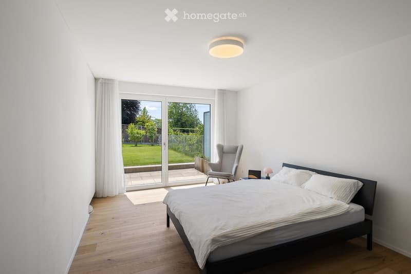 Bright bedroom with wooden flooring, large sliding glass doors leading to a garden, a bed with white bedding, a grey armchair, and a small table lamp on a nightstand.
