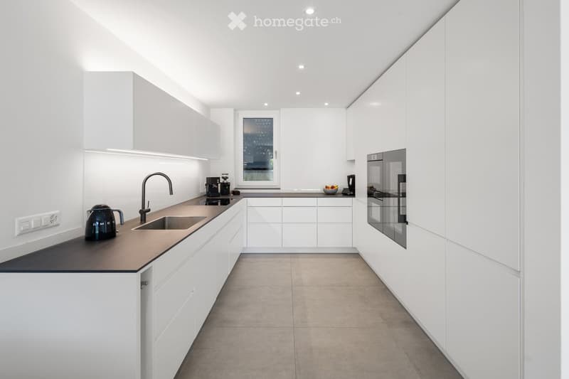 Modern, minimalist kitchen with white cabinets, dark countertops, and built-in appliances. Features a sink, faucet, coffee maker, refrigerator, kettle, and fruit bowl on the counter.