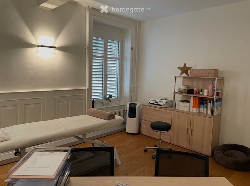 A well-lit room with a treatment table, a stool, a wooden cabinet, a shelf with books and items, a printer, a chair, a dog bed, a small plant, and a wall-mounted light fixture.