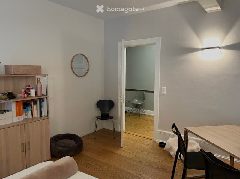 A cozy, well-lit room with wooden flooring, a small table with chairs, a shelf with books and a water bottle, a dog bed, and a mirror on the wall.