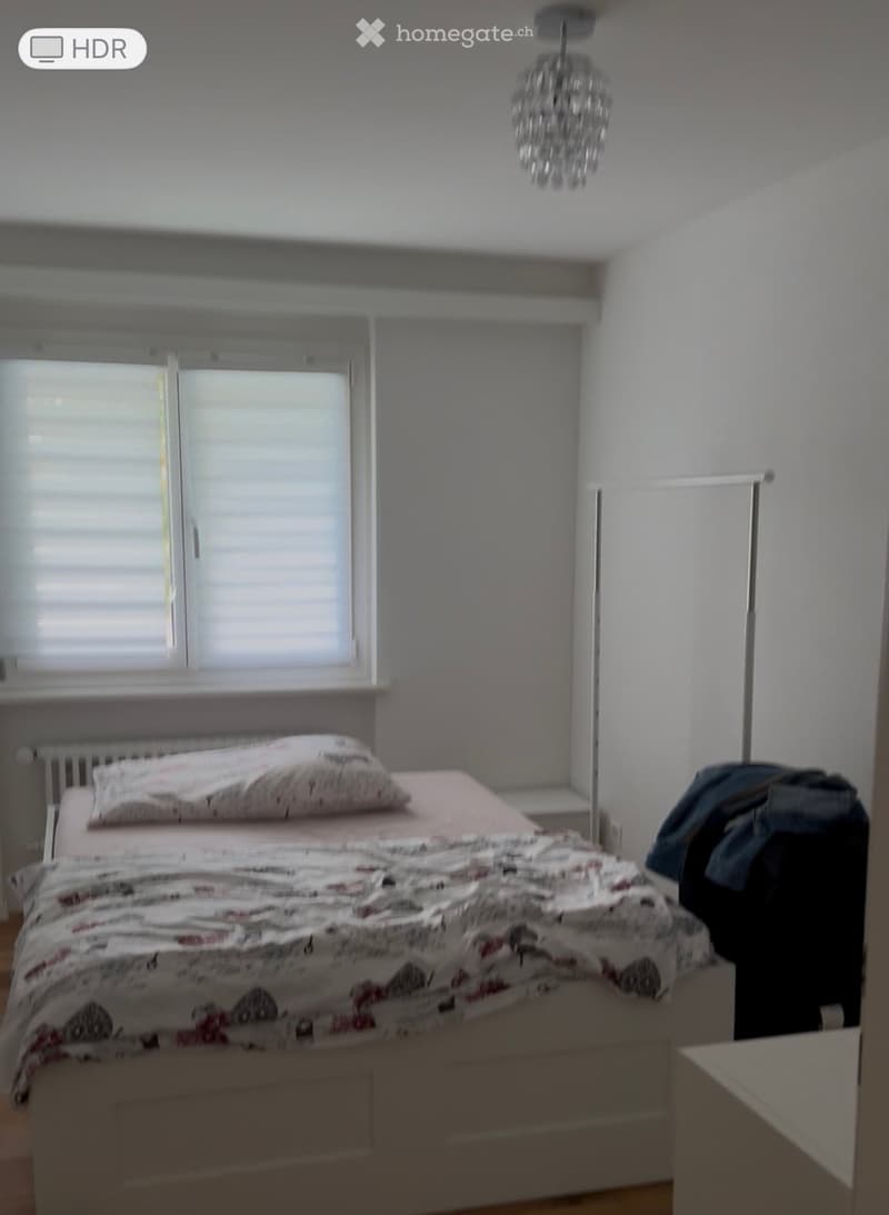 A well-lit bedroom with a bed, pillows, and a patterned blanket. The room features a window with blinds, a radiator, a chandelier, and a wardrobe with clothes.