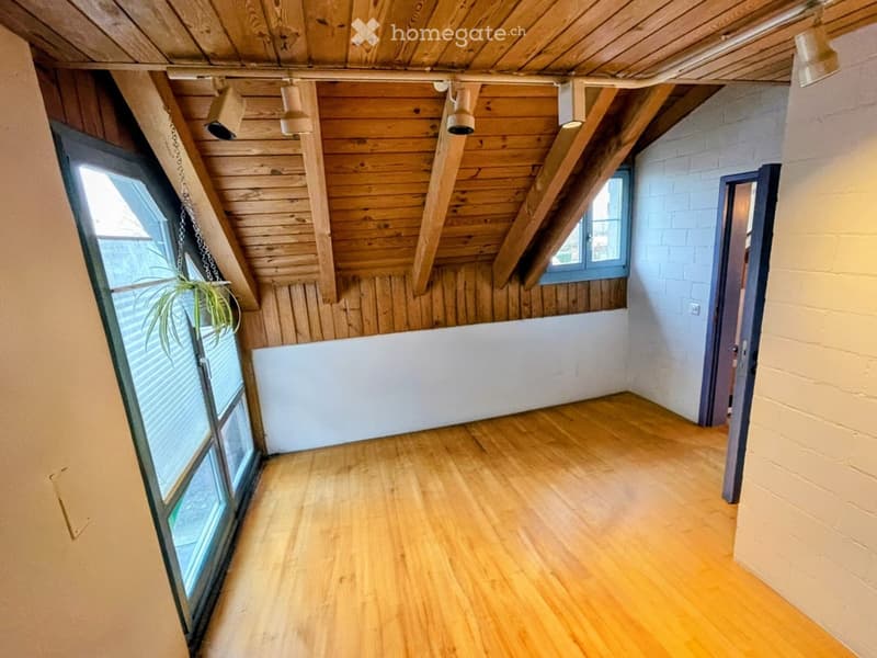Empty room with wooden flooring, exposed wooden beams on the ceiling, and large windows allowing natural light. The walls are painted white, and there is a small plant in a hanging basket near one window.