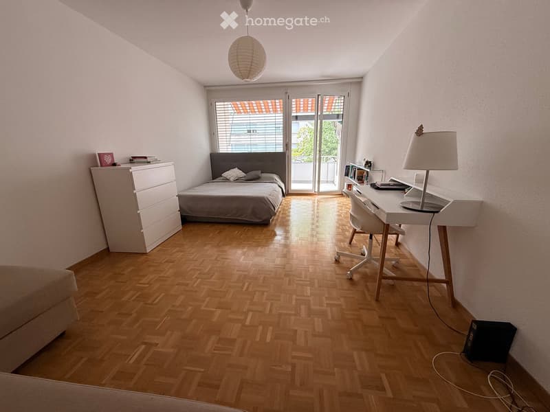 Bright bedroom with wooden flooring, a double bed with grey bedding, a nightstand, a desk with a chair, and a hanging lamp.