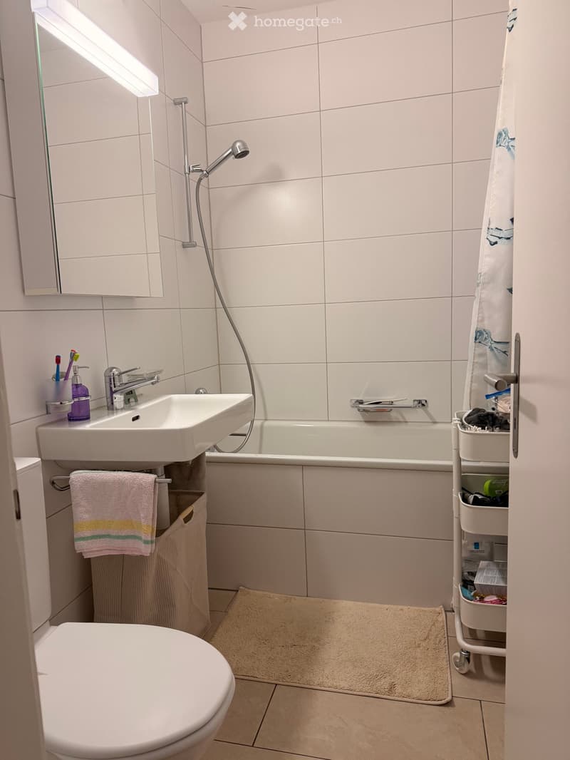 Modern bathroom with white tiles, a sink, toilet, bathtub, and a towel on a rack. A small cart holds various items.