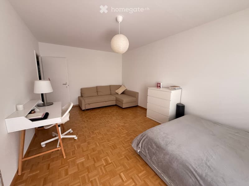 A furnished room with a bed, sofa, desk, chair, and a lamp. The room features wooden flooring and white walls.
