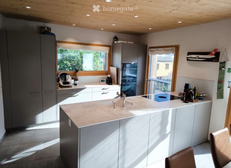 Modern kitchen with a central island, white cabinets, and a large window. Features include a coffee maker, a faucet, and a chair near the island.
