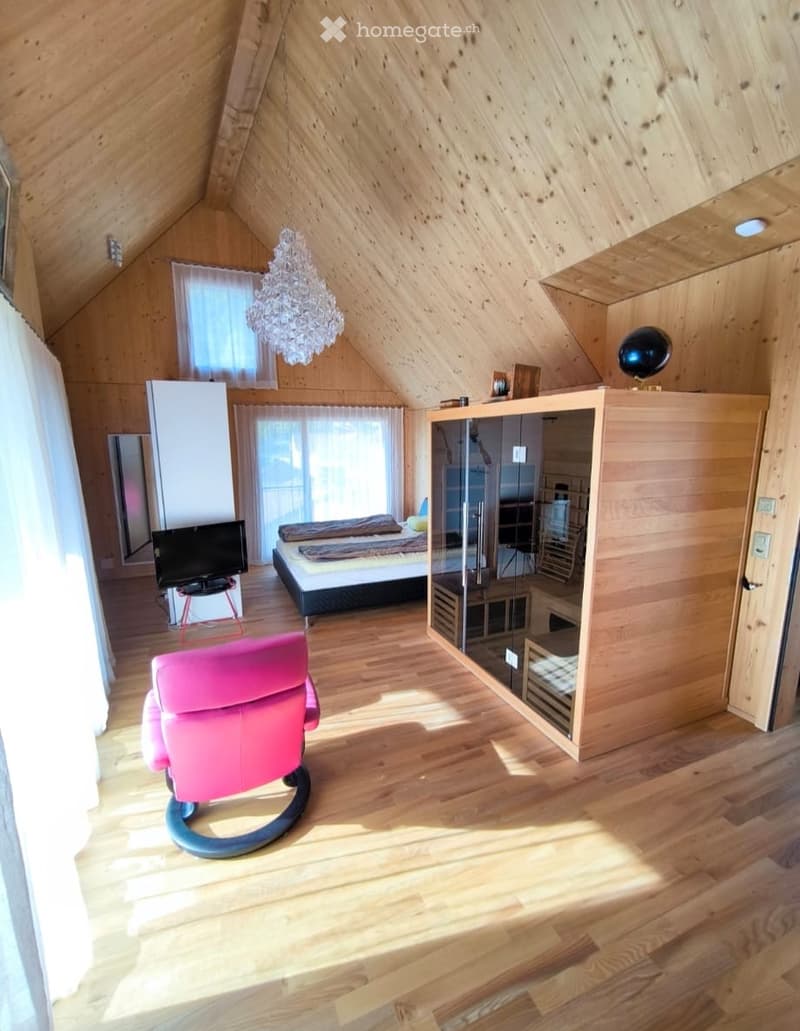 Cozy attic bedroom with wooden walls and floors, featuring a bed, pink chair, TV, chandelier, and a mirrored closet.