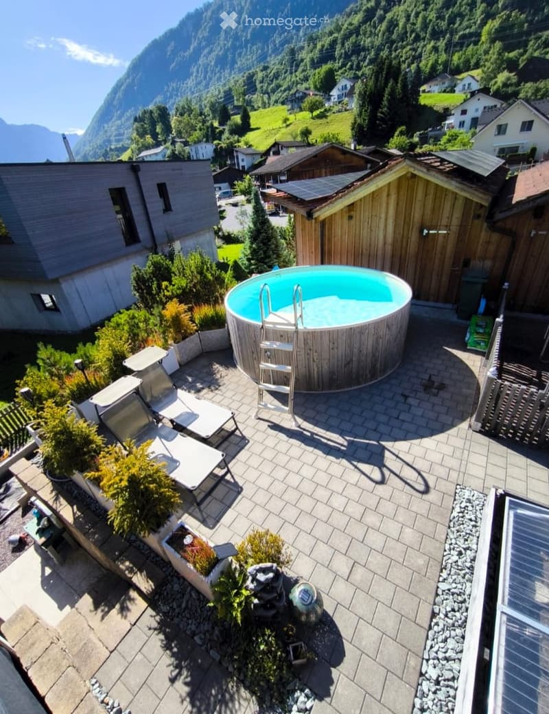 A serene outdoor area featuring a round wooden hot tub with a ladder, surrounded by lounge chairs, small plants, and a scenic mountain backdrop.