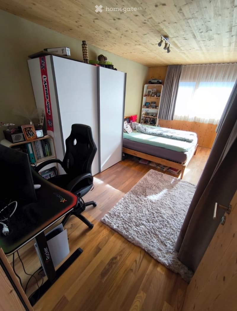 Cozy bedroom with wooden floors, slanted wooden ceiling, bed with bedding, desk with chair, bookshelf, and wardrobe.