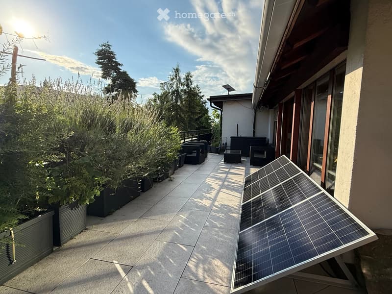 Balcony with tiled floor, several potted plants, outdoor seating, and a solar panel on the ground.