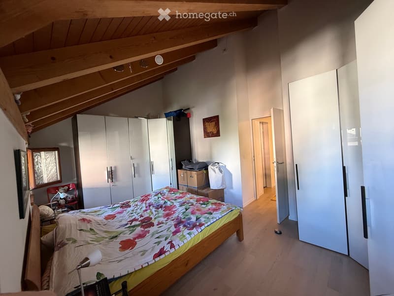 Bedroom with wooden slanted ceiling, bed with floral bedding, wardrobe, chair, lamp, and mirror. Some boxes and bags are visible near the wardrobe.
