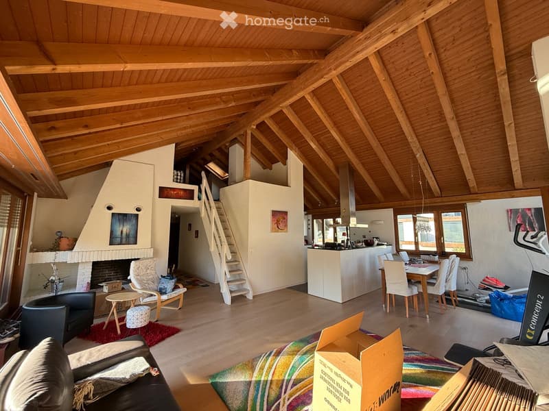 Open-plan living and dining area with exposed wooden beams, a fireplace, a staircase leading to an upper level, and a modern kitchen area with white cabinets and a counter.