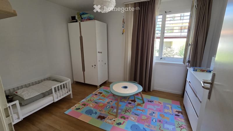A well-lit children's bedroom with a crib, a small round table, a colorful play mat, and a wardrobe. The room features wooden flooring and large windows with curtains.