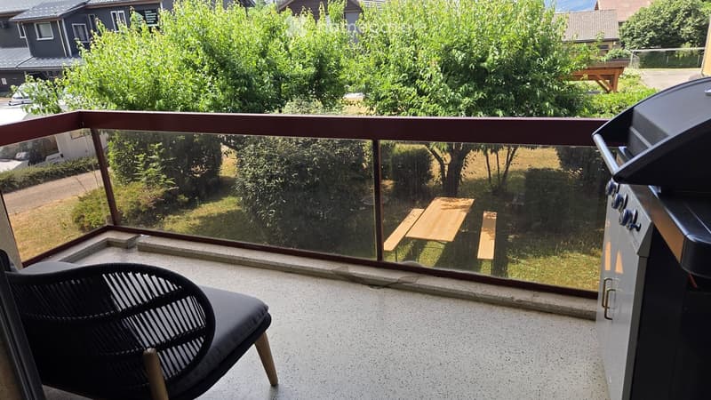 Balcony with a modern black chair, a glass railing, and a view of a green garden with a wooden table and benches.