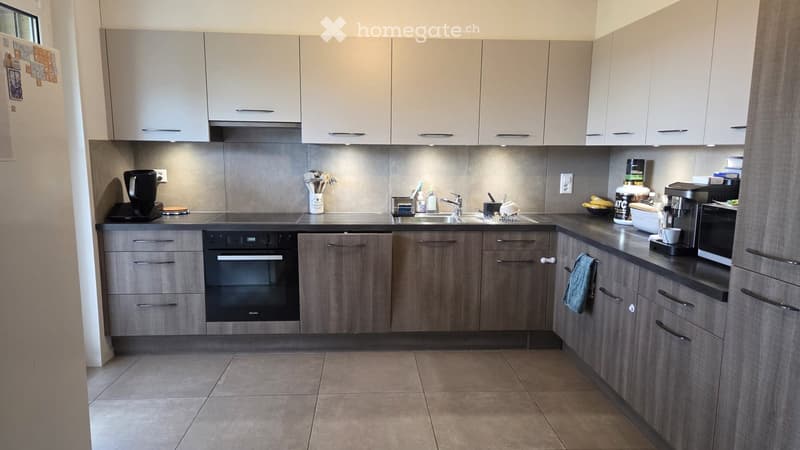 Modern kitchen with light grey cabinets, stainless steel appliances, and a tiled floor. Features an oven, microwave, coffee maker, and various kitchen items on the counter.