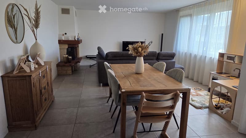 A well-lit living and dining area with a grey sofa, wooden dining table with chairs, fireplace, decorative vases, and a small desk area.