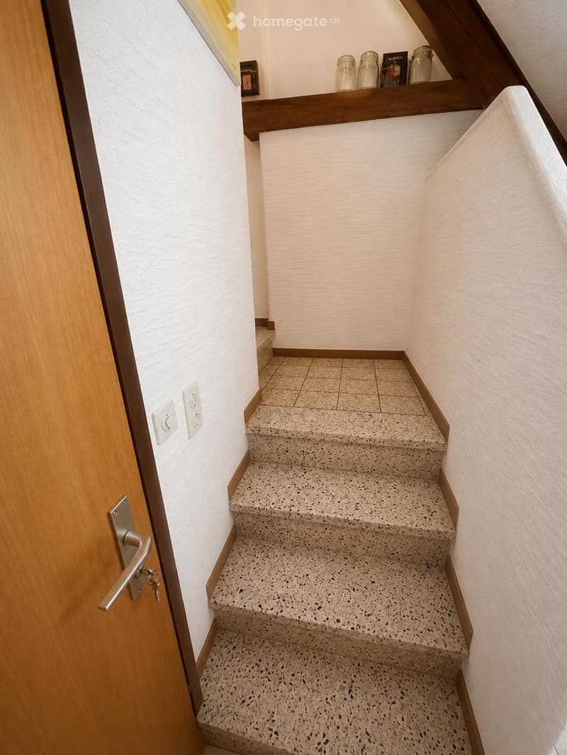 A narrow staircase with concrete steps and white walls, featuring a wooden handrail and a small shelf with jars and decorative items at the top.