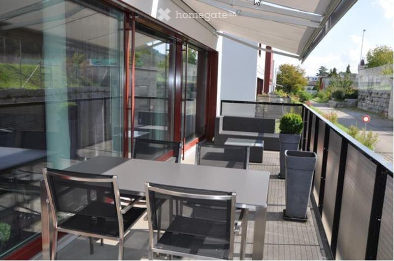 Modern balcony with glass railings, outdoor furniture including a table and chairs, and a small seating area with a couch and side table.
