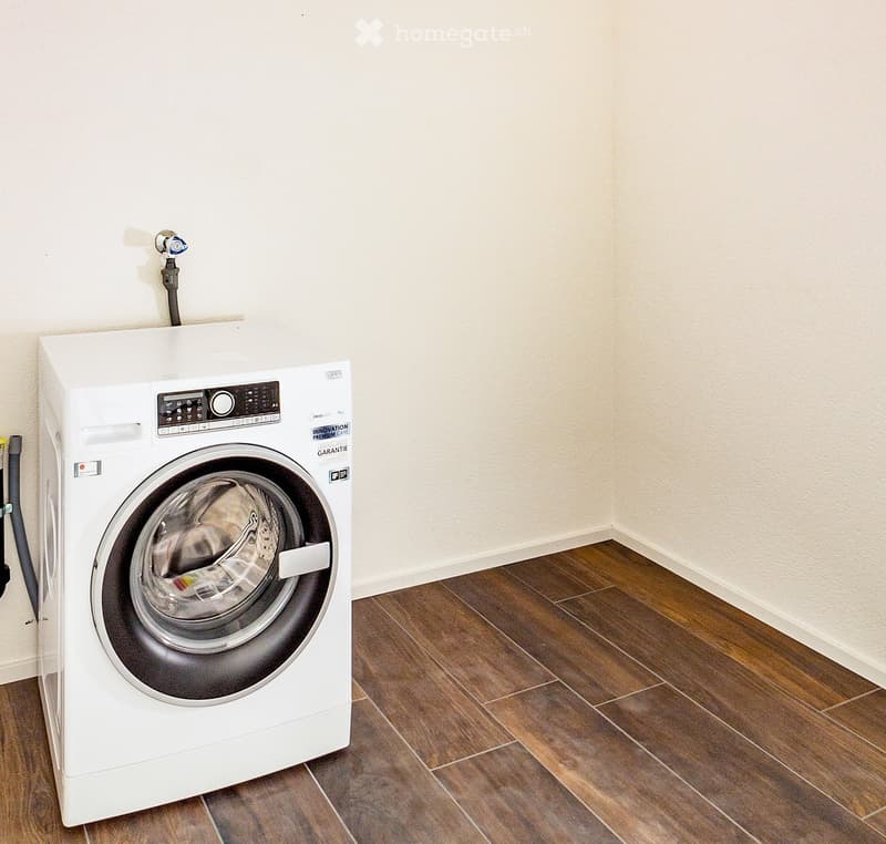 A modern, white washing machine is placed on a wooden floor in a clean, unfurnished room with light-colored walls.