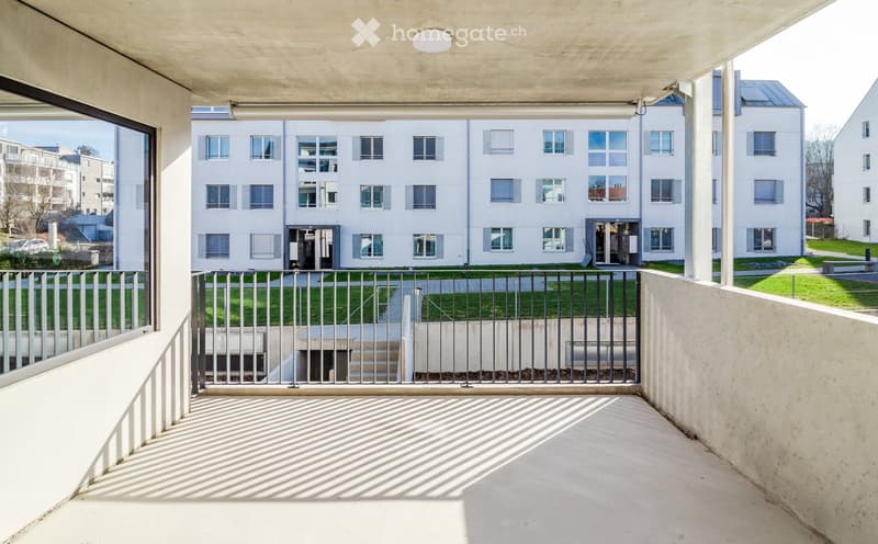 Balcony with metal railing, overlooking a modern multi-story building with numerous windows and a landscaped area below.