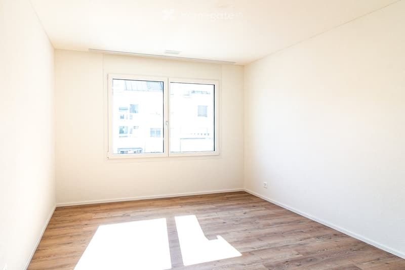 Empty room with wooden flooring, white walls, and large windows offering a view of neighboring buildings.