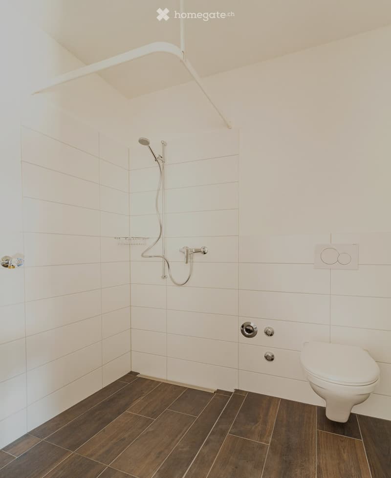 Modern bathroom with white tiled walls, dark wooden flooring, a toilet, a showerhead, and a faucet.
