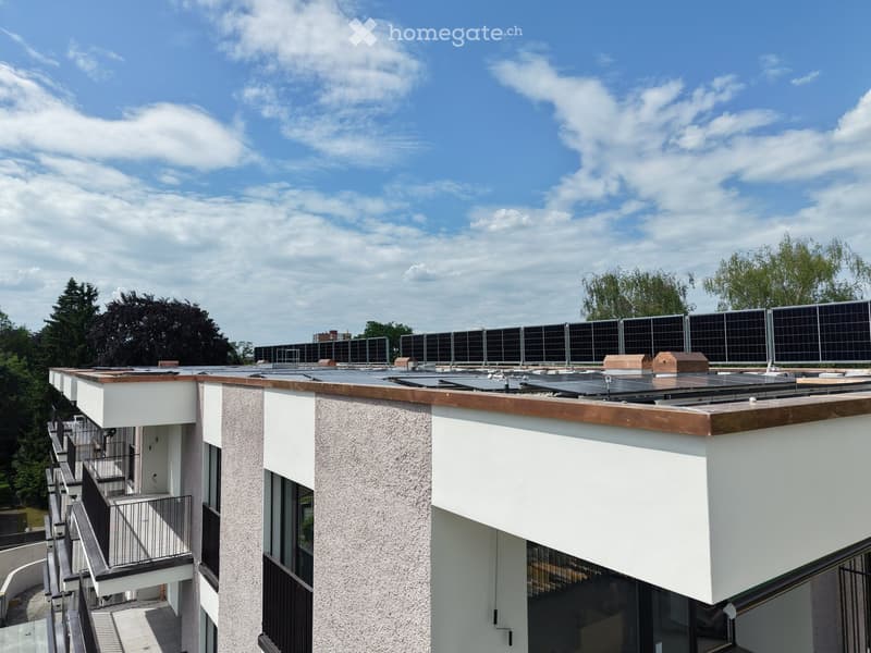 Modern multi-story building with solar panels on the roof, white and grey exterior, balconies, and surrounding greenery.