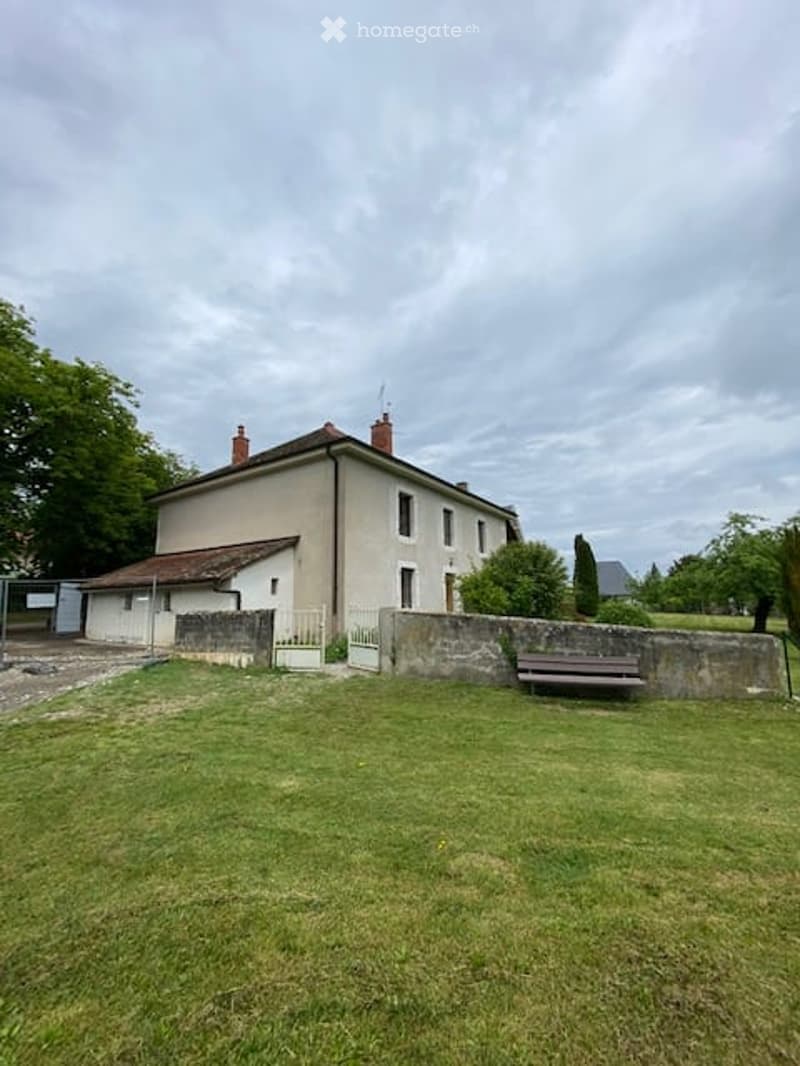 A two-story house with a beige facade, red-tiled roof, and chimneys. The house is surrounded by a low stone wall and a grassy lawn, with a bench placed near the wall. There are trees and a gate visible in the background.