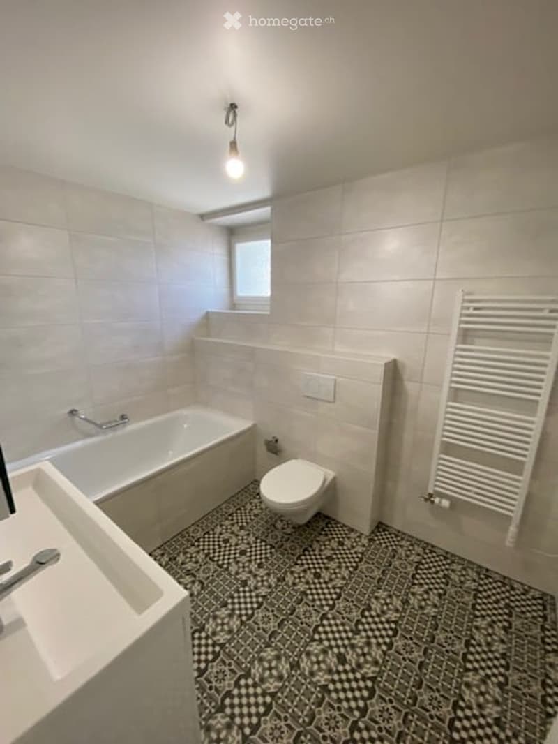 Modern bathroom with white tiles, a bathtub, toilet, sink, and a white radiator. The floor features a black and white checkered tile design.