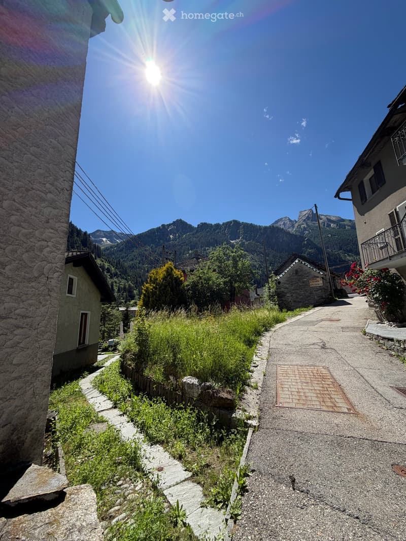 A narrow, sunlit pathway flanked by stone walls and greenery, leading up a hillside with houses and mountains in the background.