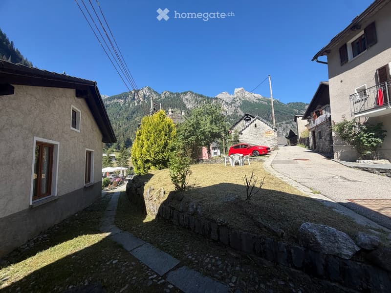A narrow stone-paved pathway leads to a small grassy area with a few trees and a red car parked nearby. The background features a stone house, a balcony with a red cloth, and distant mountain peaks under a clear blue sky.