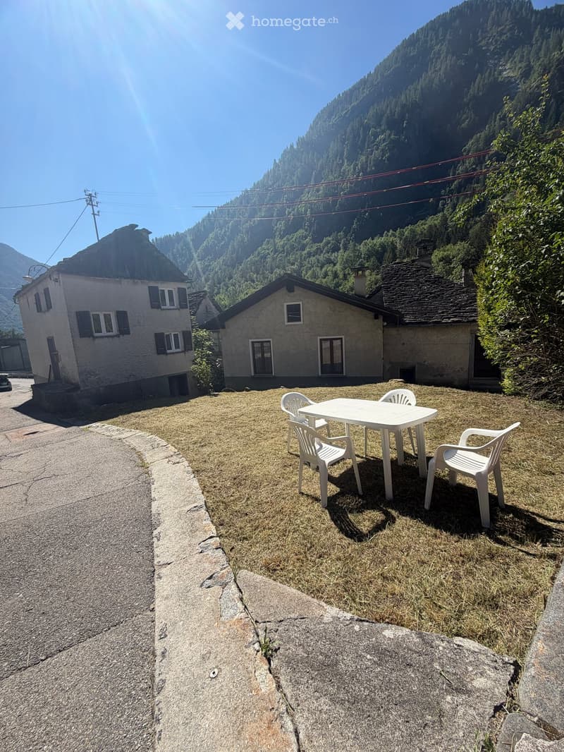 Outdoor seating area with a white plastic table and four chairs on a grassy patch, surrounded by houses and a mountainous backdrop.