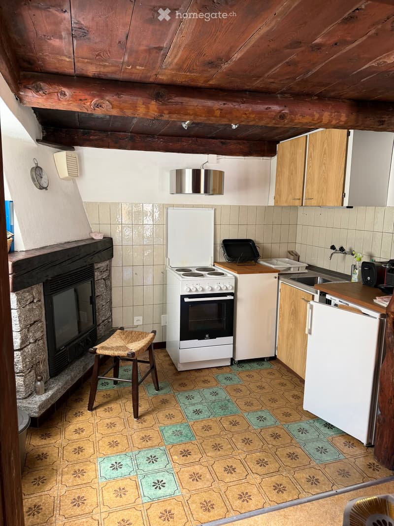 Cozy kitchen with wooden beams, fireplace, stove, refrigerator, sink, and wooden stool.