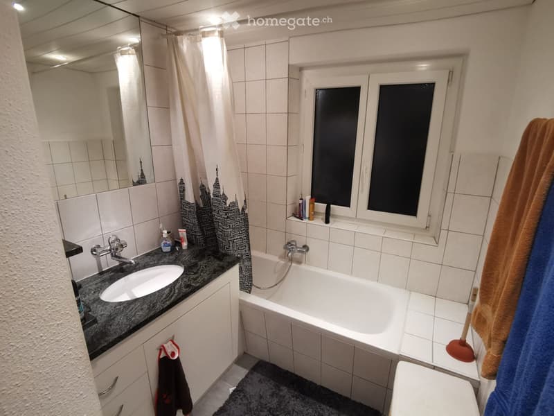 Bathroom with white tiles, a bathtub, a sink with a mirror, and a window with frosted glass. Towels and a toilet are visible.