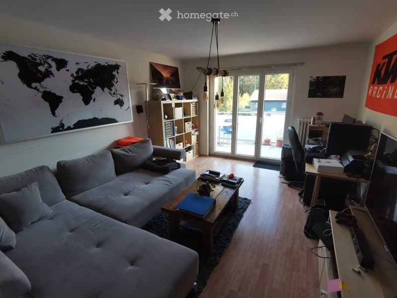 A modern living room with a grey sectional sofa, wooden coffee table, laptop on a desk, and a bookshelf. Large windows and a sliding glass door lead to a balcony.
