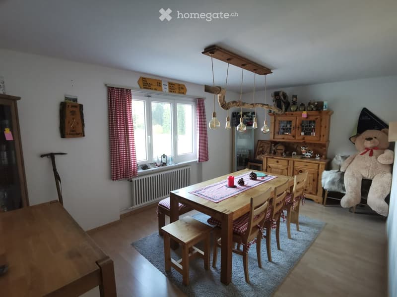 Brightly lit dining area with wooden table and chairs, a large window with checkered curtains, and a wooden cabinet with glass doors.