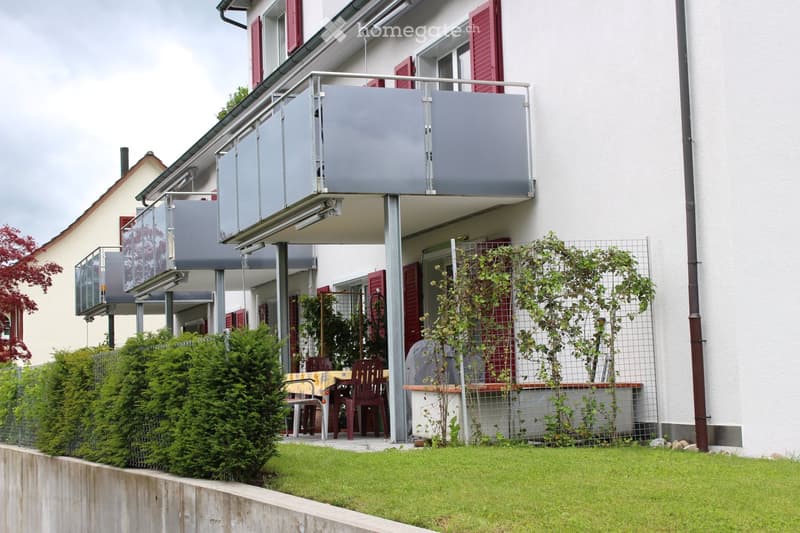 Balcony and garden area with a table and chairs, surrounded by greenery and a modern house with red shutters.
