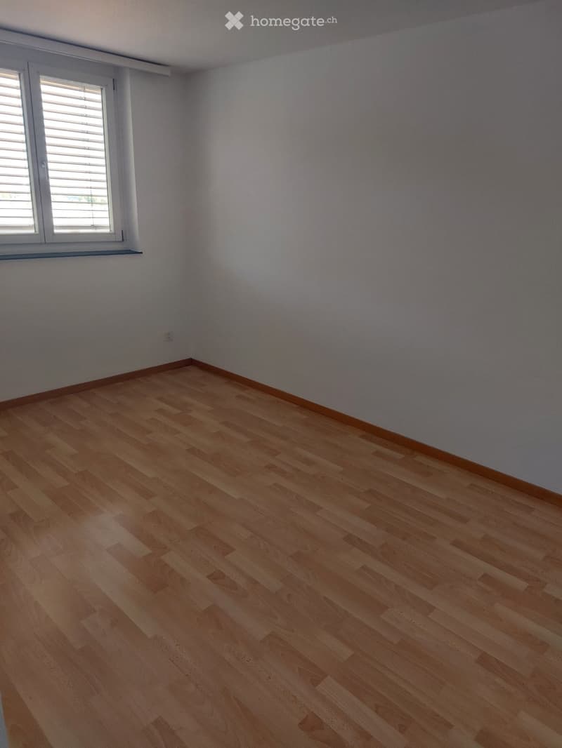 Empty room with light wooden flooring and white walls, featuring a window with blinds on the left side.