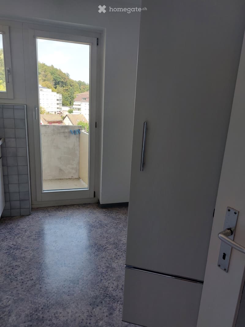 Empty room with a tiled wall, a door, and a large window leading to a balcony with a view of buildings and trees.