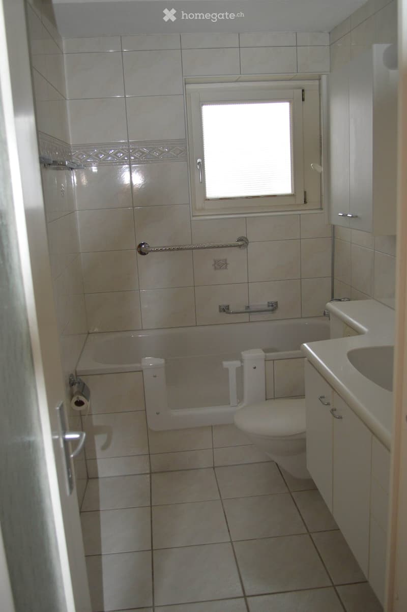 A small bathroom with tiled walls and floor, featuring a toilet, a sink with a cabinet, a bathtub, and a window above the bathtub. Toilet paper is visible on a holder next to the toilet.