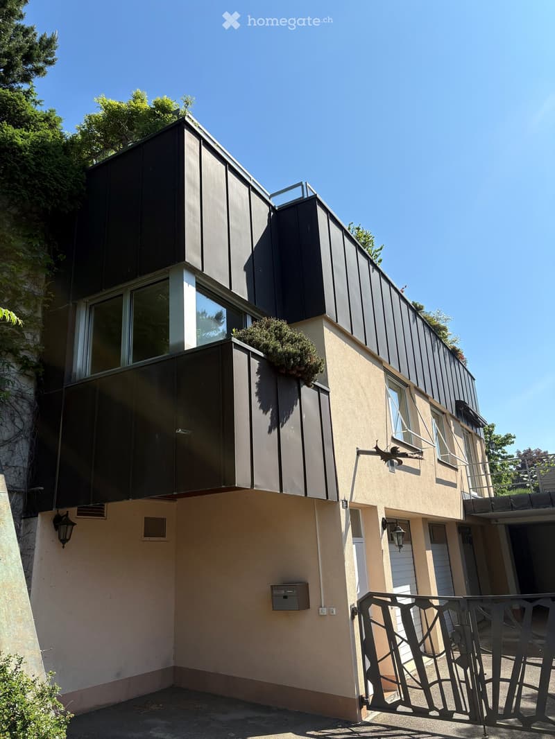 Modern two-story building with a combination of dark metal siding and light beige walls, featuring large windows, a small balcony with greenery, and a gated entrance.